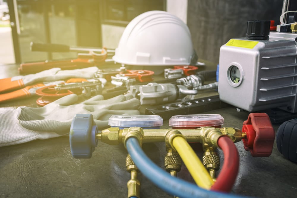 HVAC equipment on a workbench including a manifold gauge set, vacuum pump, hard hat, and gloves.