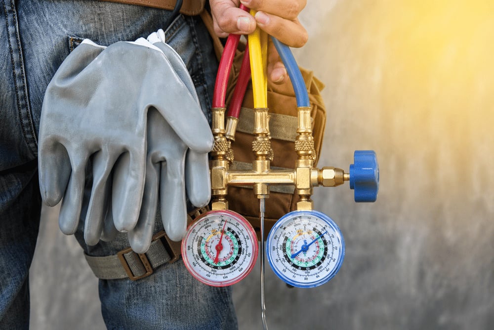 A close-up of a technician holding an HVAC manifold gauge set, with work gloves tucked into his back pocket.